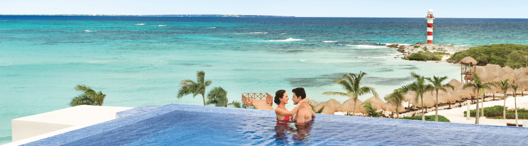 A woman and a man in an infinity pool that looks over the sandy beaches of a resort and the clear blue ocean with a lighthouse in the background