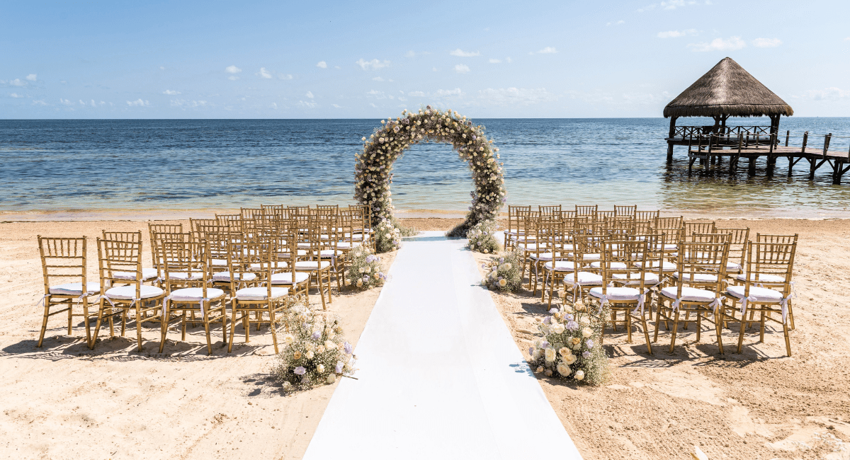 View of a destination wedding on a beach with a white walkway for the bride and a circular arch for the couple to stand at for the ceremony with two sections of chairs for the guests overlooking the ocean with a bungalow in the background