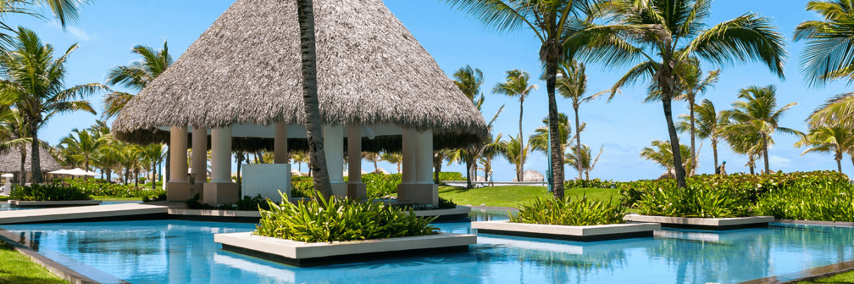 View of a pool area at an all inclusive resort with a bamboo thatched palm-leaf roof building and palm trees in the background 